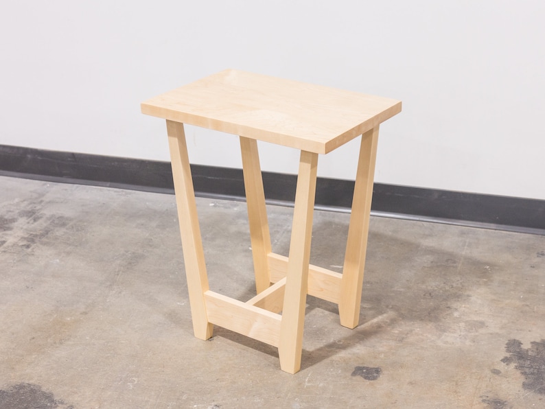 A side table placed in a living room with a plant and coffee mug sat on the table top. The side table is made from light maple hardwood and is crafted in Scandinavian minimalist modern design.