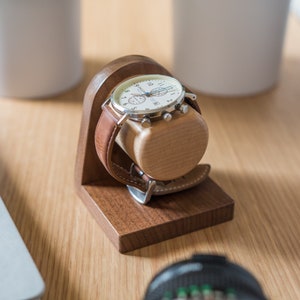 One watch rests on a smooth maple dowel which rests on a strong walnut base. Set on a desk with notebook and camera.