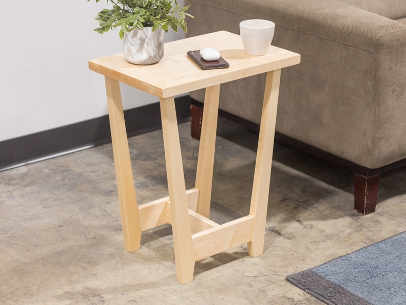 A side table placed in a living room with a plant and coffee mug sat on the table top. The side table is made from light maple hardwood and is crafted in Scandinavian minimalist modern design.