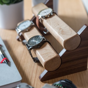 Three watches rest on a smooth maple dowel which rests on a strong walnut base. Set on a desk with notebook and camera.