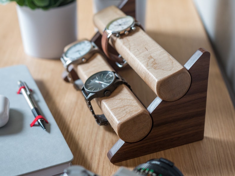 Three watches rest on a smooth maple dowel which rests on a strong walnut base. Set on a desk with notebook and camera.