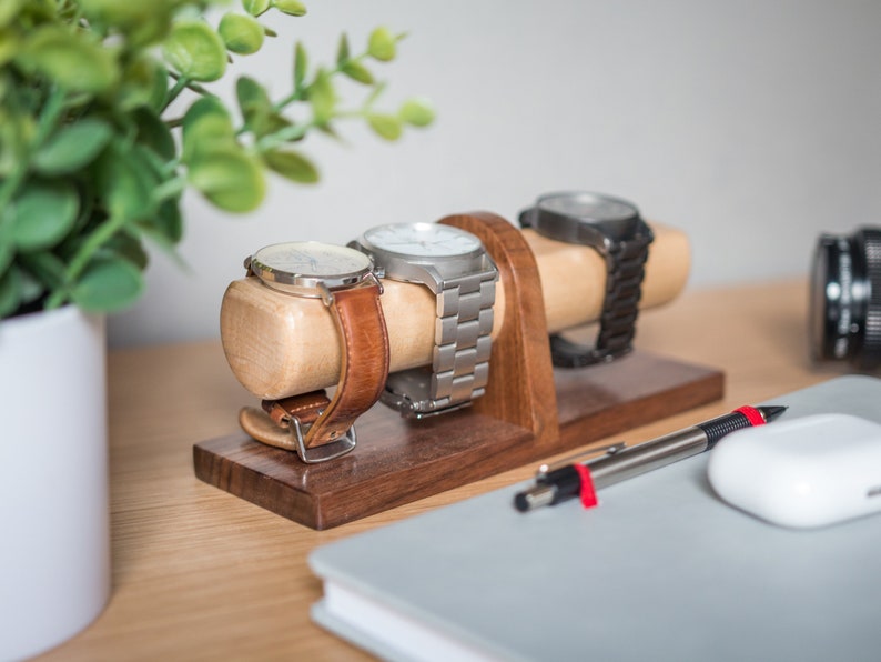 Watch display holder rests on a table, made of a long, wooden block base with one round dowel sticking out horizontally, enough space to hold four watches.