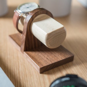 Two watches rest on a smooth maple dowel which rests on a strong walnut base. Set on a desk with notebook and camera. Light and warm wood contrasts well with mechanical metal watch.