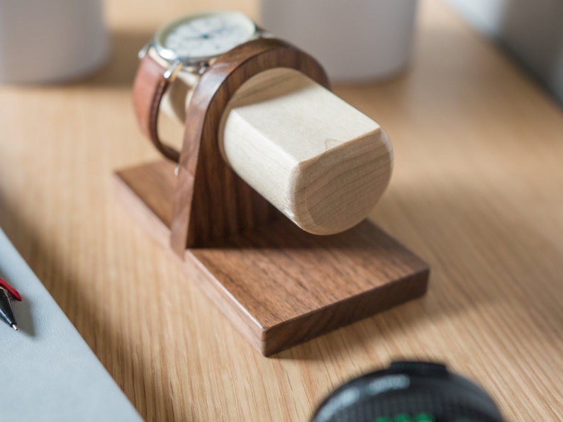 Two watches rest on a smooth maple dowel which rests on a strong walnut base. Set on a desk with notebook and camera. Light and warm wood contrasts well with mechanical metal watch.