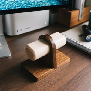 May include: A wooden watch stand with a light brown cylindrical base and a dark brown base. The stand is on a dark brown wooden surface.