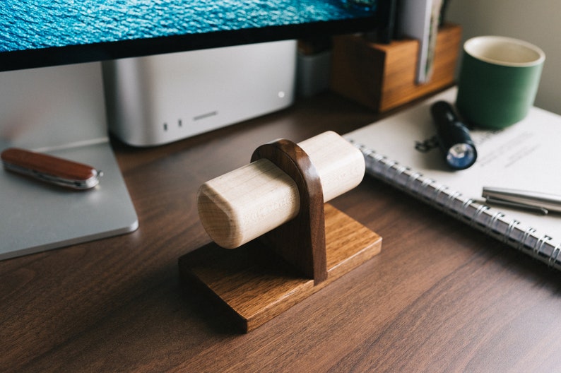 May include: A wooden watch stand with a light brown cylindrical base and a dark brown base. The stand is on a dark brown wooden surface.