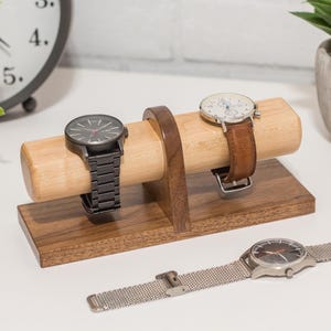 May include: A wooden watch stand with two watches on it. The stand is made of walnut wood and has a light brown finish. The watches are both analog and have metal bands.