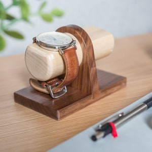 Two watches rest on a smooth maple dowel which rests on a strong walnut base. Set on a desk with notebook and camera. Light and warm wood contrasts well with mechanical metal watch.