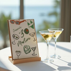 May include: A wooden sign with the text "Jessica's 30 Martini" and illustrations of martini glasses, olives, sunglasses, and a swimsuit. Two martini glasses with olives are on a white table in front of the sign.