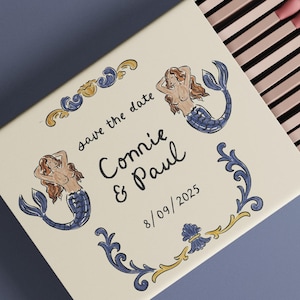 May include: A decorative matchbox with a cream-colored surface, featuring illustrations of mermaids and ornate blue and gold accents. The text "save the date" and names "Connie & Paul" are displayed, along with the date 8/09/2025. The matches have pink tips.