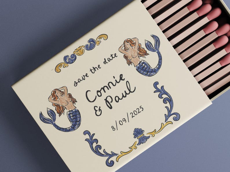 May include: A decorative matchbox with a cream-colored surface, featuring illustrations of mermaids and ornate blue and gold accents. The text "save the date" and names "Connie & Paul" are displayed, along with the date 8/09/2025. The matches have pink tips.