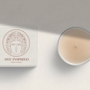 K&ouml;nnte beinhalten: Eine wei&szlig;e Box mit einer braunen Linienzeichnung eines Frauenkopfes mit einem Lorbeerkranz. Der Text "AWE INSPIRED AROMA THERAPY" befindet sich unter der Zeichnung. Neben der Box befindet sich eine durchsichtige Glaskerze mit beigefarbenem Wachs.