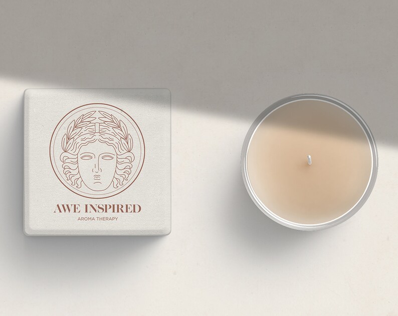 K&ouml;nnte beinhalten: Eine wei&szlig;e Box mit einer braunen Linienzeichnung eines Frauenkopfes mit einem Lorbeerkranz. Der Text "AWE INSPIRED AROMA THERAPY" befindet sich unter der Zeichnung. Neben der Box befindet sich eine durchsichtige Glaskerze mit beigefarbenem Wachs.