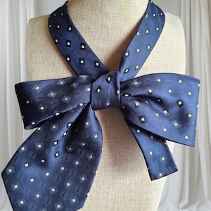 May include: A navy blue bow tie with a repeating pattern of white and grey squares and diamonds. The bow tie is displayed on a mannequin head. The fabric has a subtle sheen.