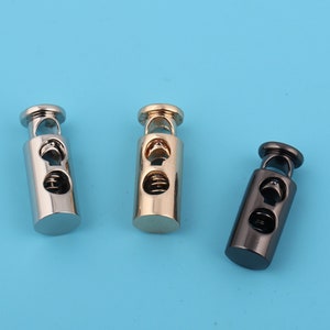 Może przedstawiać: Trzy metalowe stopery do sznurka w kolorze srebrnym, złotym i czarnym. Każdy stoper ma mechanizm sprężynowy do regulacji długości sznurka.