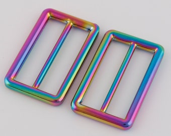 Op de afbeelding: Twee regenboogkleurige metalen schuifgespen. De gespen zijn rechthoekig met een enkele, gecentreerde, horizontale gleuf.