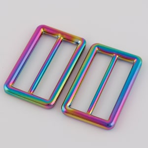 Puede incluir: Dos hebillas deslizantes de metal iridiscente. Las hebillas son rectangulares con una sola ranura horizontal centrada.
