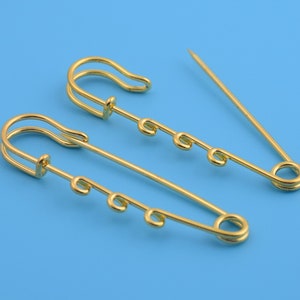 May include: Two gold safety pins with multiple loops on the pin. The pins are on a blue background.