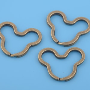 May include: Three bronze-colored key rings in a stylized, three-lobed shape, resembling a cartoon character's head. The key rings are open, ready to attach to keys or other items. The background is a solid light blue.