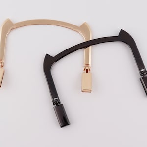 Puede incluir: Dos asas de bolso, una dorada y otra negra, con un diseño de bisagra. Las asas son de metal y tienen una forma rectangular en los extremos.