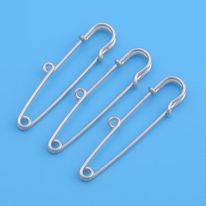 Op de afbeelding: Drie zilveren veiligheidsspelden, elk met een gesloten lus aan de bovenkant en een puntige punt. De spelden zijn in een rij gerangschikt, met de bovenste speld iets verschoven.