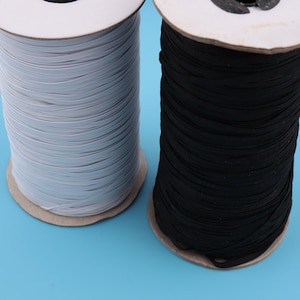 May include: Two spools of elastic cord, one white and one black, are wound around cardboard spools. The spools are sitting on a blue surface.
