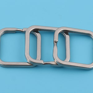 Op de afbeelding: Drie zilveren vierkante sleutelringen van metaal, op elkaar gestapeld.