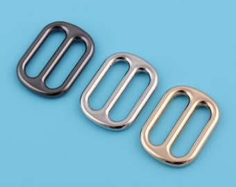 May include: Three metal rectangle sliders in gunmetal, silver, and gold colors. Each slider has two parallel bars and a central opening. The sliders are arranged diagonally on a light blue background, suitable for various crafting projects.