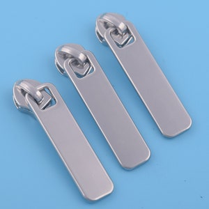 Puede incluir: Tres tiradores de cremallera plateados con un diseño rectangular y un lazo superior redondeado. Están dispuestos sobre un fondo azul claro, creando una estética limpia y moderna. Probablemente para la reparación de ropa o bolsos.