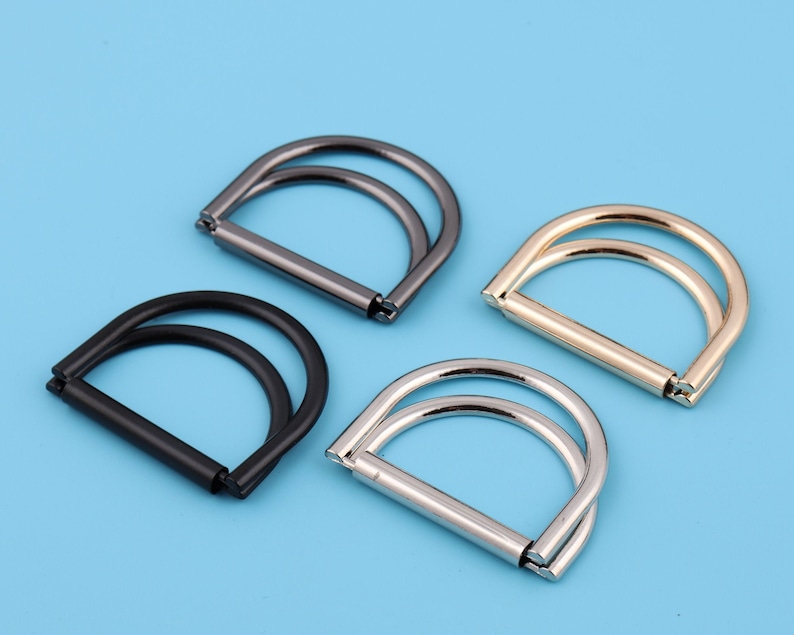 May include: Four metal D-rings in different finishes: black, silver, gold, and nickel. The rings are double-layered and have a smooth, polished finish.