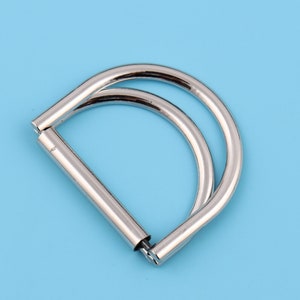 May include: A silver metal D-ring with a double loop. The ring is designed for attaching to bags, purses, or other items.