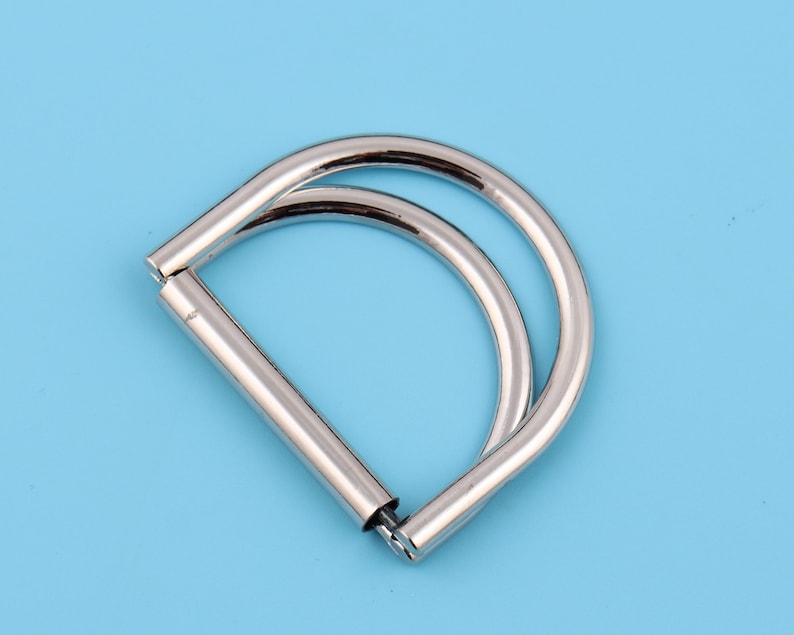May include: A silver metal D-ring with a double loop. The ring is designed for attaching to bags, purses, or other items.