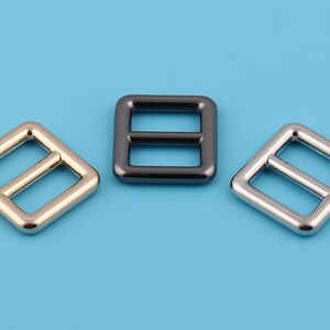 Pu&ograve; includere: Tre fibbie quadrate in metallo con una barra centrale. Le fibbie sono dorate, argentate e nere.