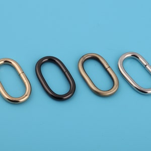 Puede incluir: Cuatro anillos metálicos ovalados en diferentes acabados: dorado, negro, bronce y plateado. Los anillos están dispuestos en una fila sobre un fondo azul.