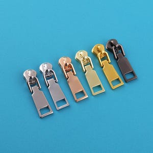 May include: A collection of six metal zipper pulls in various colors, including silver, bronze, gold, and black. Each pull has a rectangular shape with a square opening at the bottom and a rounded top with a loop for attachment. The pulls are arranged on a solid blue background.