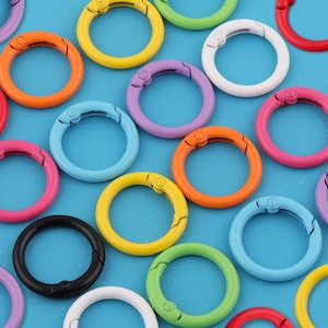 Pode incluir: Uma coleção de mosquetões redondos de metal coloridos em várias tonalidades, incluindo vermelho, laranja, amarelo, verde, azul, roxo, rosa, preto e branco. Os clipes estão dispostos sobre um fundo azul sólido, mostrando suas cores vibrantes e design funcional.