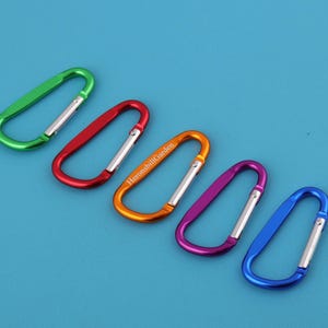 以下が含まれることがあります： 緑、赤、オレンジ、紫、青の5色のカラビナクリップが、青色の背景に斜めに配置されています。オレンジ色のクリップには「HeronsbillGarden」の文字がプリントされています。各クリップには銀色のゲートが付いています。