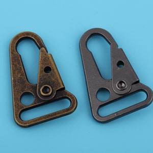 Op de afbeelding: Twee metalen trigger-snaphaken, een in antiek goud en een in zwart. Beide haken hebben een driehoekige vorm met een afgeronde opening en een D-ring.