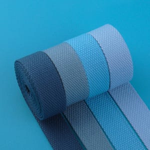 May include: A roll of blue webbing in various shades, from deep navy to light sky blue, against a solid blue background. The webbing has a textured, woven appearance, suggesting durability and a range of potential uses.
