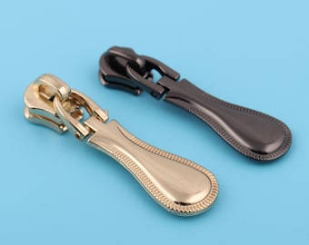 May include: Two zipper pulls, one gold and one black, with a decorative design. The pulls are shaped like a teardrop with a textured edge.