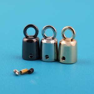 May include: Three metal bell-shaped charms in black, silver, and gold, each with a loop at the top and a small hole near the base. Small screws are also visible. The charms are on a blue background.