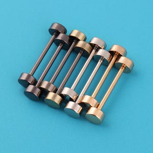 May include: A selection of six barbell stud earrings in different metallic finishes, including dark brown, gold, and silver. The earrings are arranged diagonally against a bright blue background.