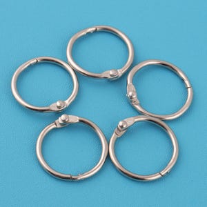 Puede incluir: Cinco anillas plateadas sobre un fondo azul. Cada anilla es circular con una abertura articulada y un pequeño cierre plateado. Las anillas son de un material metálico brillante.