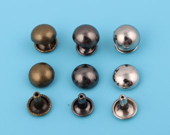 Könnte beinhalten: Sortiment runder Metallnieten in drei Farben: Antikmessing, Schwarz und Silber. Die Nieten haben eine gewölbte Oberseite und eine flache Unterseite mit einem Stift zum Befestigen an Stoff oder Leder.