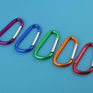 Può includere: Cinque moschettoni colorati, viola, blu, verde, arancione e rosso, disposti diagonalmente su sfondo blu. Il moschettone rosso ha il testo "HeronsbillGarden". Ogni moschettone ha un cancello argentato.