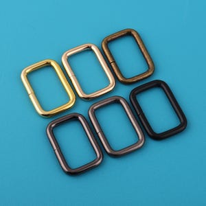 Può includere: Sei anelli rettangolari in metallo in varie finiture: oro, ottone, bronzo, nero e canna di fucile. Gli anelli sono disposti su uno sfondo turchese. Questi anelli sono probabilmente utilizzati per la fabbricazione di borse o altri progetti artigianali.