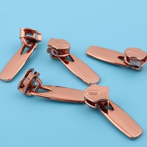 May include: Four rose gold zipper pulls. The zipper pulls are made of metal and have a smooth, shiny finish.