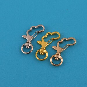 May include: Three metal swivel clasps in silver, gold, and rose gold. Each clasp features a heart-shaped top, a spring-loaded clip, and a circular ring at the base. The clasps are arranged against a solid blue background.