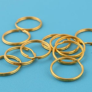 Może przedstawiać: Kolekcja złotych kółek do kluczy z podziałem, rozrzuconych na jasnoniebieskiej powierzchni. Pierścienie są wykonane z metalu i mają podzieloną konstrukcję ułatwiającą mocowanie. Obraz jest dobrze oświetlony, podkreślając metaliczny połysk pierścieni.