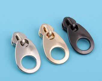 May include: Three zipper pulls in different finishes: silver, gold, and black. Each pull has a rounded oval shape with a hole in the center.